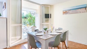 Un comedor con una mesa dispuesta para cuatro personas, un ventanal corredero que da a un patio con plantas y un cuadro con una playa en la pared.