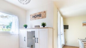 Una cocina moderna con muebles blancos, una ventana con cortinas ligeras, un letrero con la palabra 'HOME' en la pared y una pequeña planta en maceta sobre la encimera.
