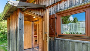 A wooden cabin with a door leading to a bedroom, a window with lace curtains, and a metal gutter on the roof.
