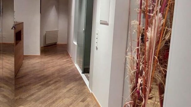 Un couloir doté d’un plancher en bois, d’une porte en verre et d’un vase rose contenant des plantes séchées.