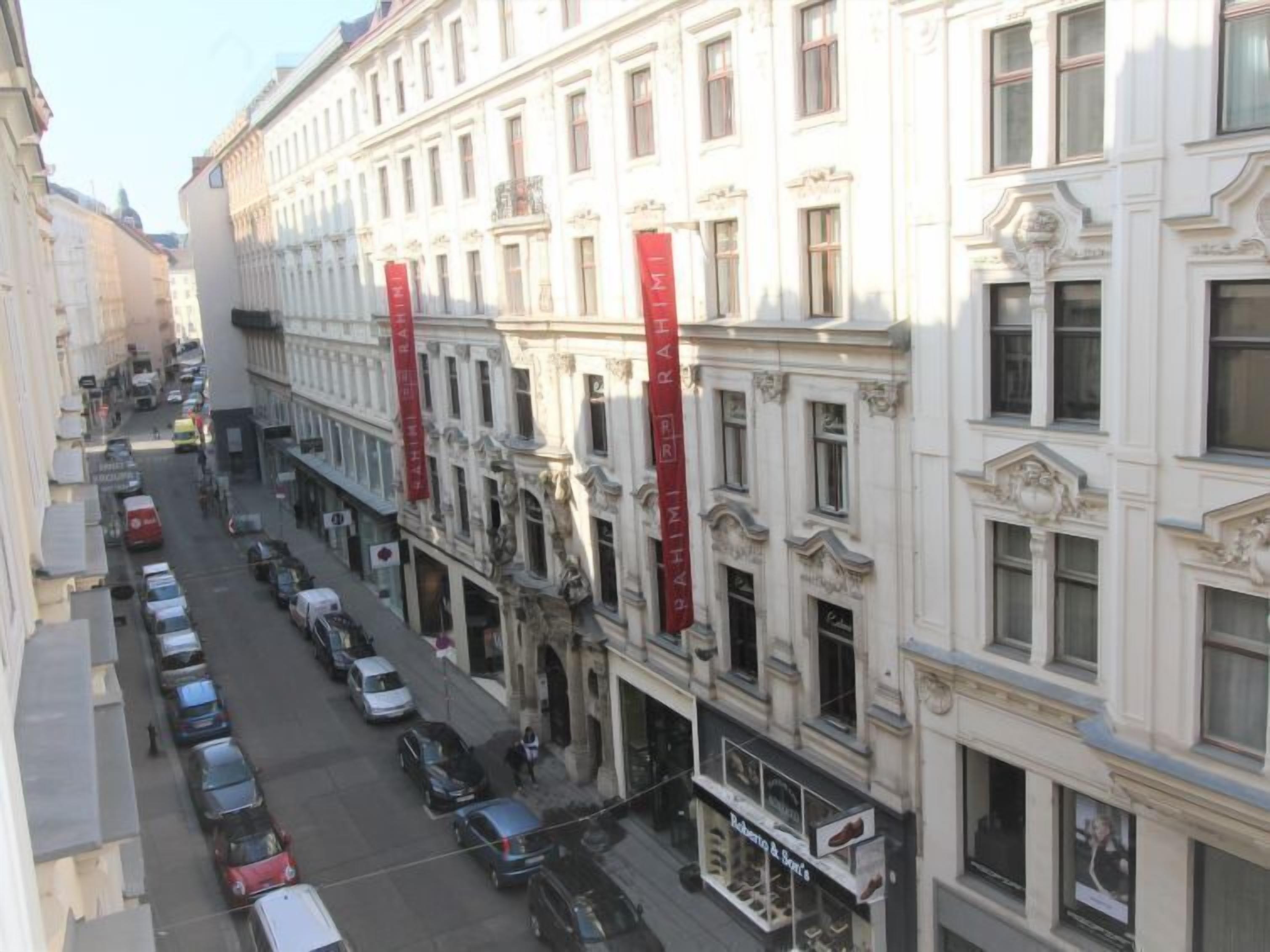 A street view of a row of historic buildings with red banners, parked cars, and a storefront with a shoe display.