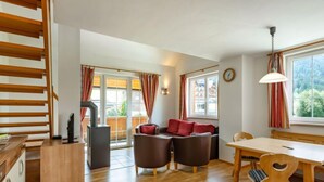 A spacious living area with a wooden staircase, a sofa set, a dining table, and a large window with curtains.