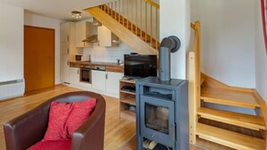 A modern living room with a brown leather sofa, a TV, and a wood-burning stove. A kitchen is visible in the background with white cabinets and a wooden staircase.
