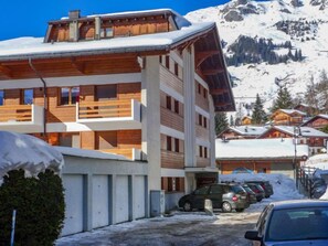 Un edificio a più piani con facciata in legno e balconi, circondato da montagne innevate e altri edifici.
