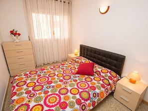 A bedroom with a bed, a dresser, a nightstand, a lamp, and a window with curtains.