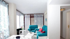 Un salon lumineux et moderne, avec une table blanche dressée pour le repas, un canapé turquoise agrémenté de coussins rouges et une grande fenêtre ornée de rideaux à motifs.