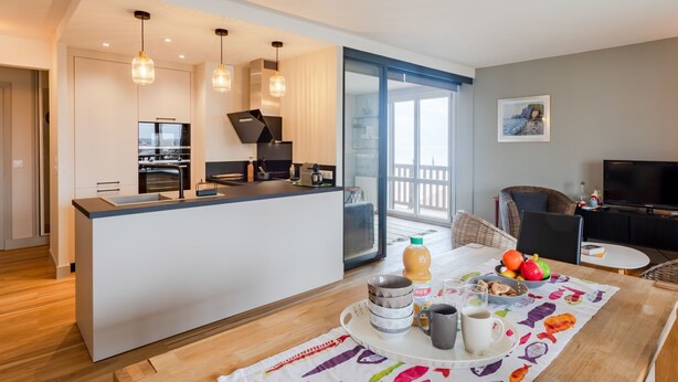 Een moderne keuken met een centraal eiland, een eethoek met een kleurrijke tafelkleed, en een woonkamer met een televisie en zitplaatsen.