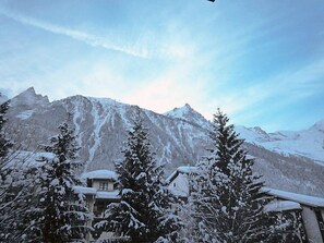 A mountain cabin with a snow-covered roof, surrounded by snow-laden trees and a backdrop of rugged mountains.