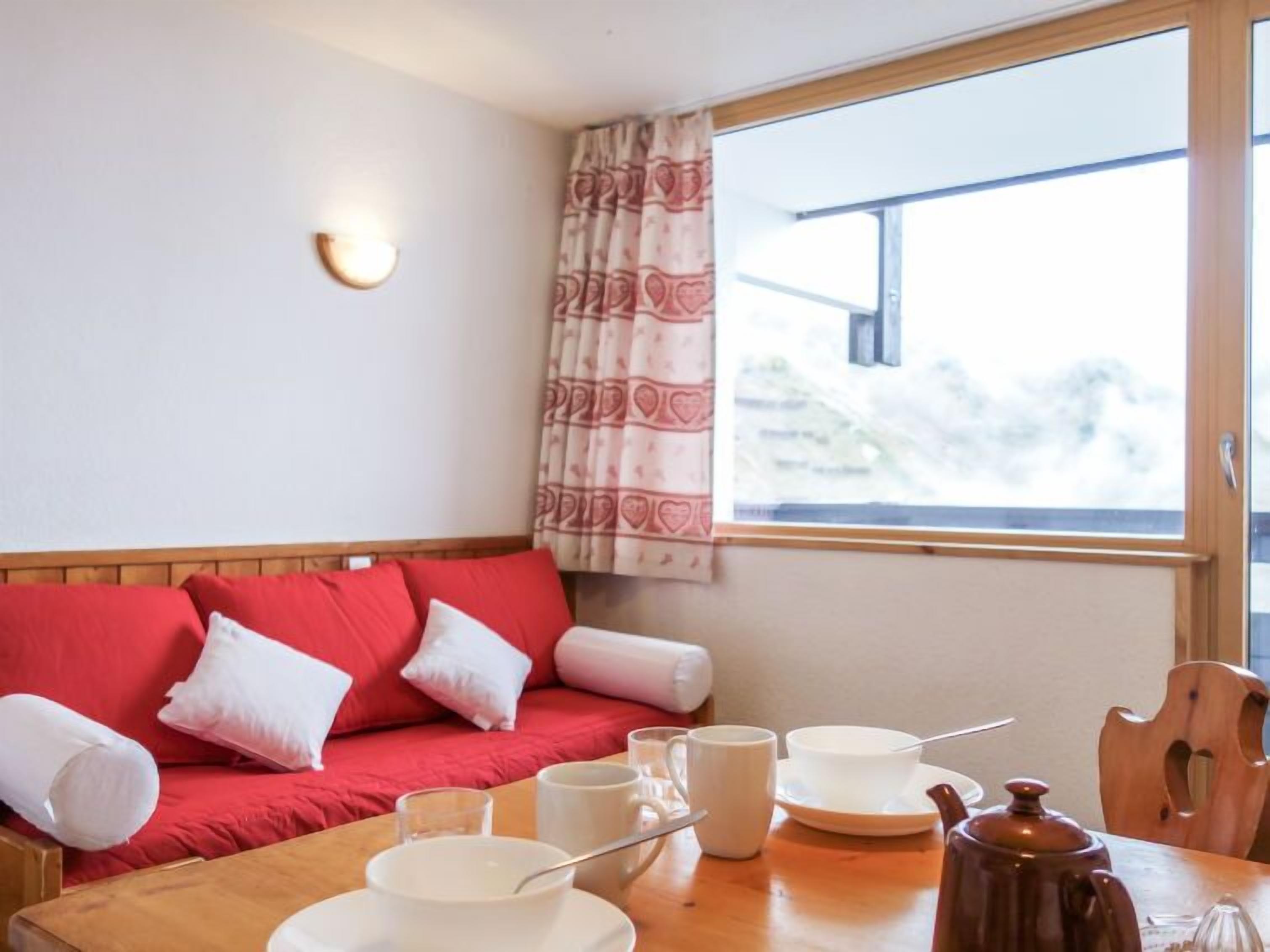 A cozy room with a red sofa, a wooden table with breakfast items, and a window with a view of trees.