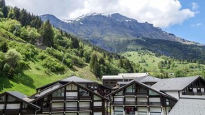 Una montagna con cime innevate e una valle boscosa. In primo piano, edifici dall'architettura tradizionale.