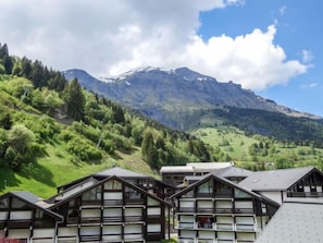 Une montagne aux sommets enneigés et une vallée boisée. Au premier plan, on aperçoit des bâtiments à l’architecture traditionnelle.