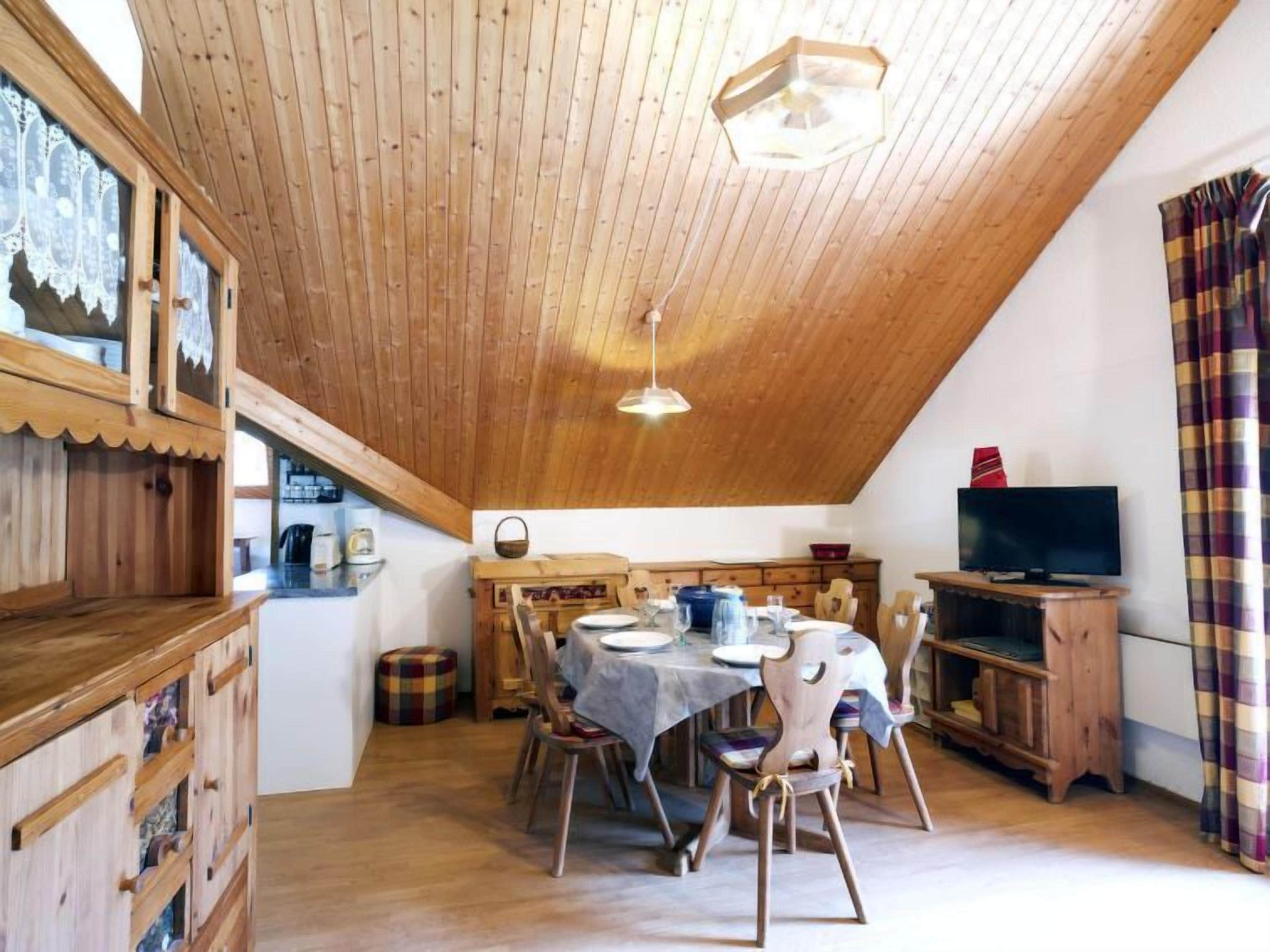 Ett mysigt rum med ett träpanelat tak, ett matbord med stolar, en tv och ett fönster med gardiner.