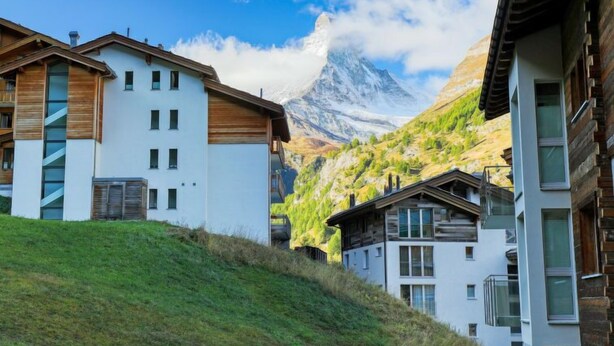 A modern building with a white facade and wooden accents, set against a backdrop of a mountain and clear sky. - Grillon by Interhome (Zermatt)