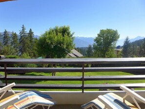 A balcony with a view of a mountain range, a grassy area, and trees.
