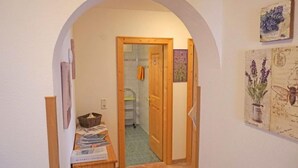 Un couloir étroit, surmonté d’une porte en arc, mène à une salle de bain. Un banc en bois, sur lequel sont posés des magazines, est adossé au mur, et une illustration botanique encadrée est accrochée sur le côté droit.