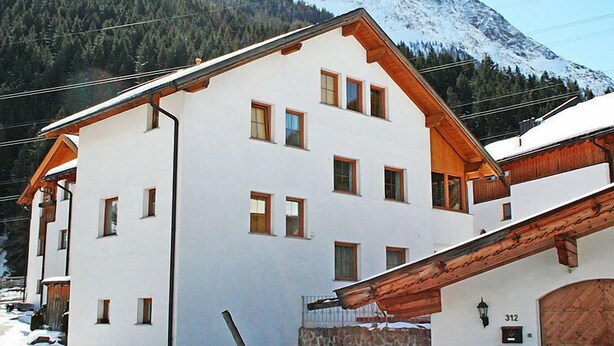 Un bâtiment à deux étages, doté d’une façade blanche et de détails en bois, se dressant sur fond de montagnes enneigées.
