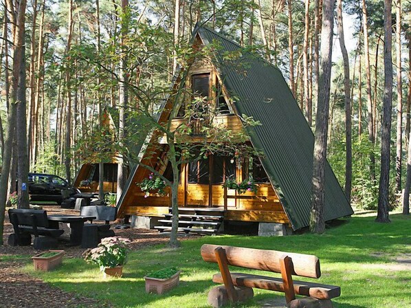 A cabin with a sloped roof, surrounded by a forest. There is a picnic table and a bench in the foreground.