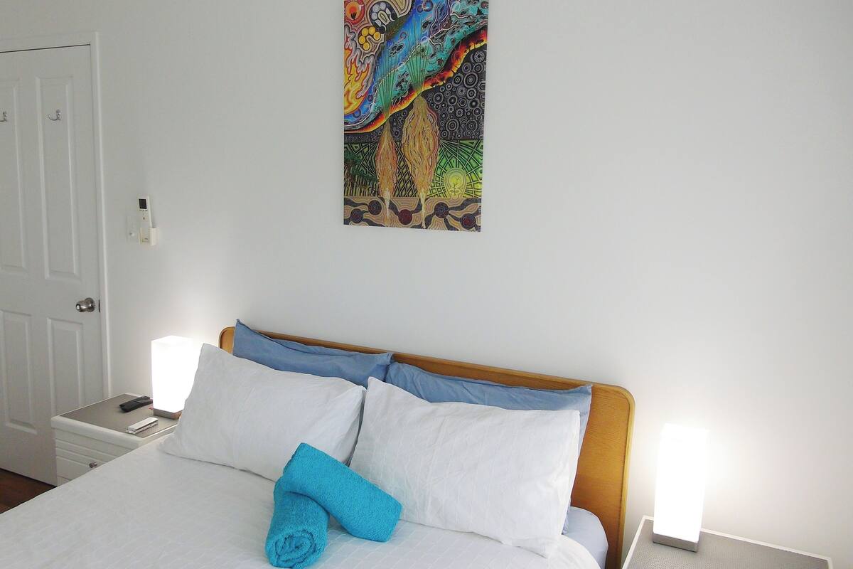 Un lit bien fait, avec une literie blanche et bleue, une serviette bleue et une peinture abstraite aux couleurs vives accrochée au mur.