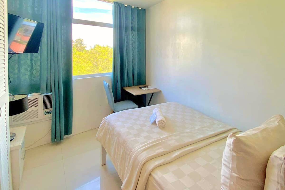 Habitación de hotel con cama, escritorio, silla, televisor y ventana con cortinas.