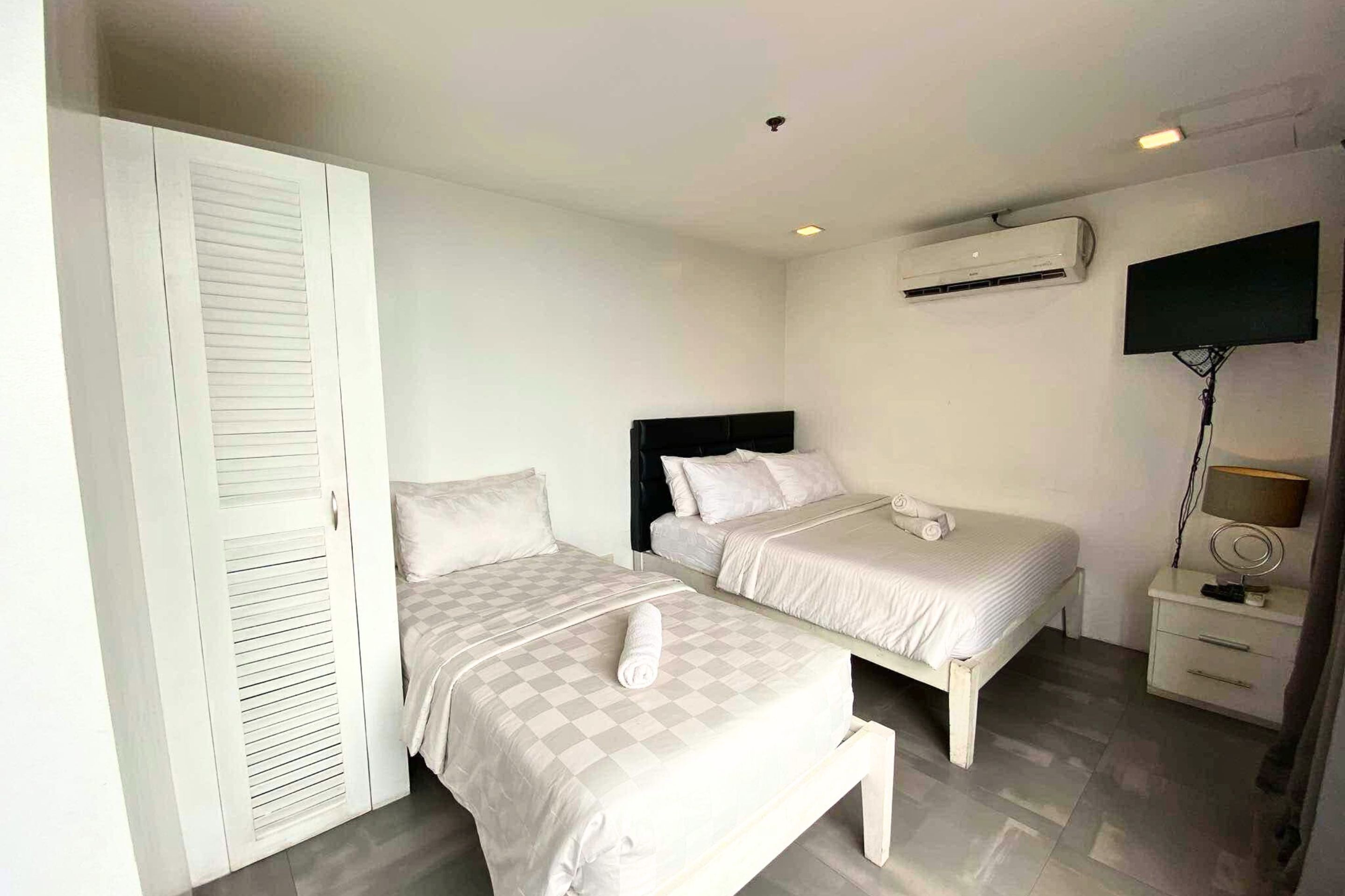 A bedroom with two beds, a wardrobe, an air conditioning unit, a TV, and a bedside table with a lamp.
