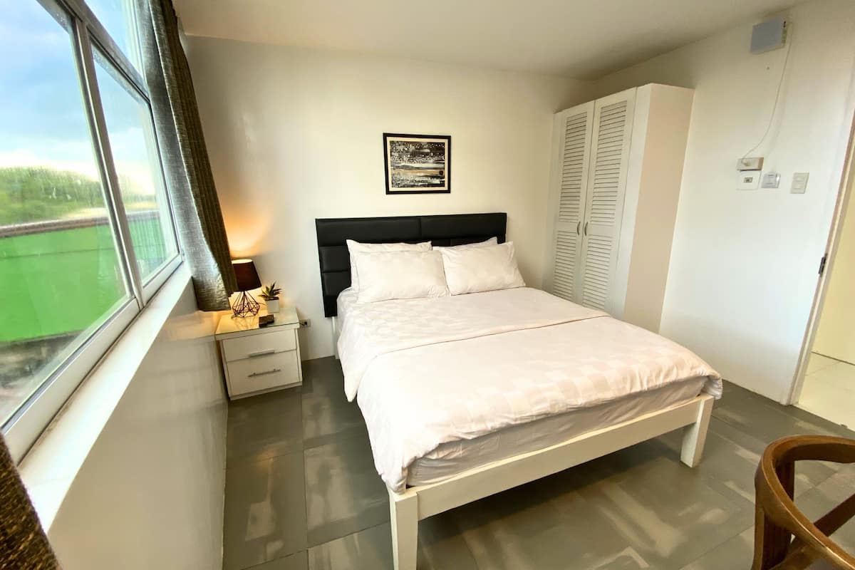 Un dormitorio con cama, mesita de noche, ventana, armario y un cuadro en la pared.