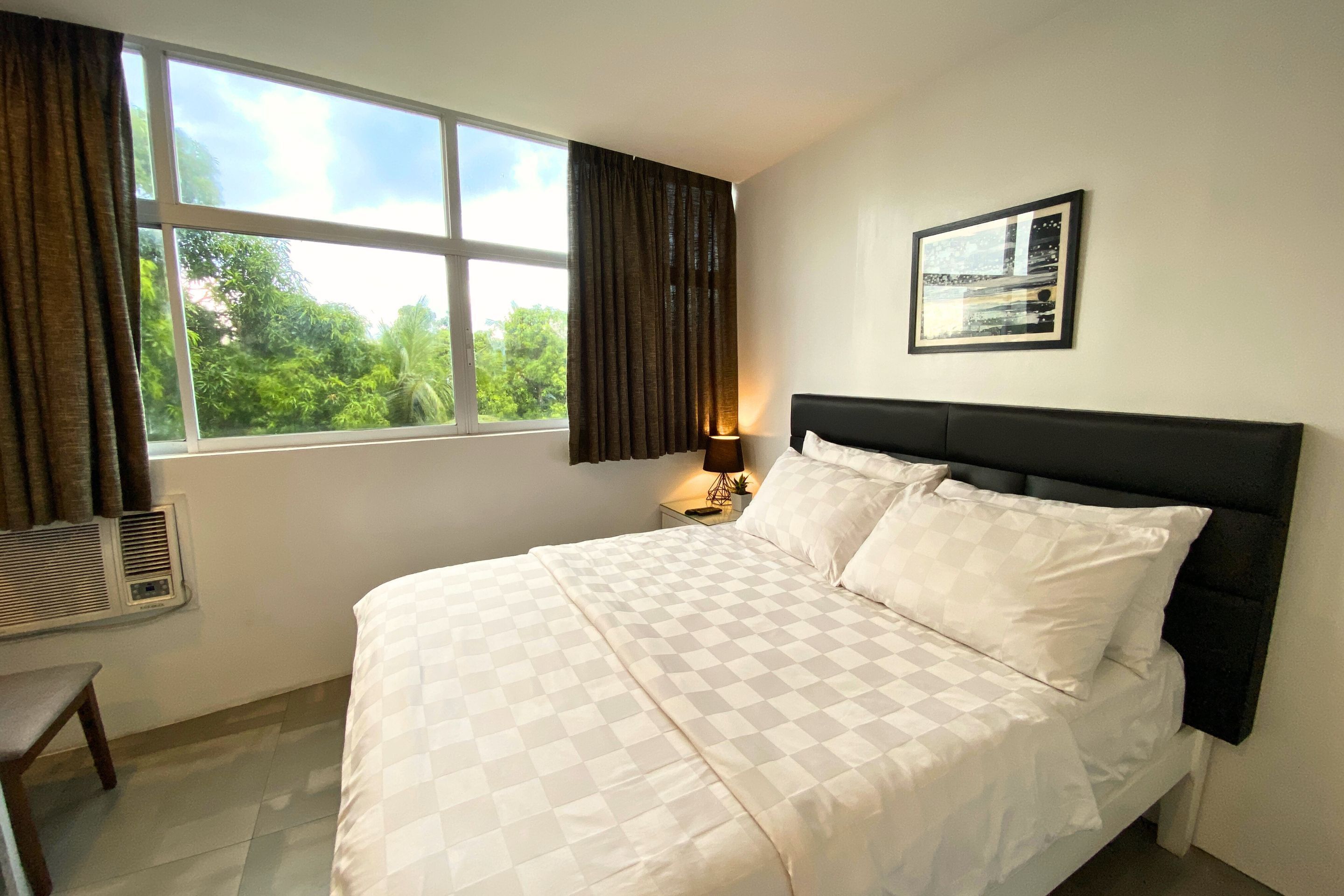 A hotel room with a large bed, a bedside table with a lamp, a window with curtains, and a framed picture on the wall.