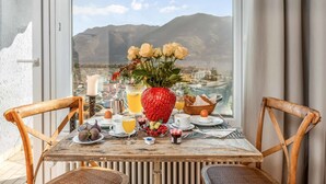 Une table de petit-déjeuner avec vue sur les montagnes et la ville, agrémentée d’un vase de fleurs, de fruits et d’un panier de pain.