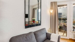 Un salon moderne avec un canapé gris, un grand miroir et une vue sur le paysage urbain, visible à travers une porte-fenêtre coulissante.
