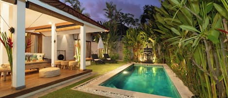 A pool area with a clear pool, surrounded by tropical plants and a covered seating area with a wooden deck.