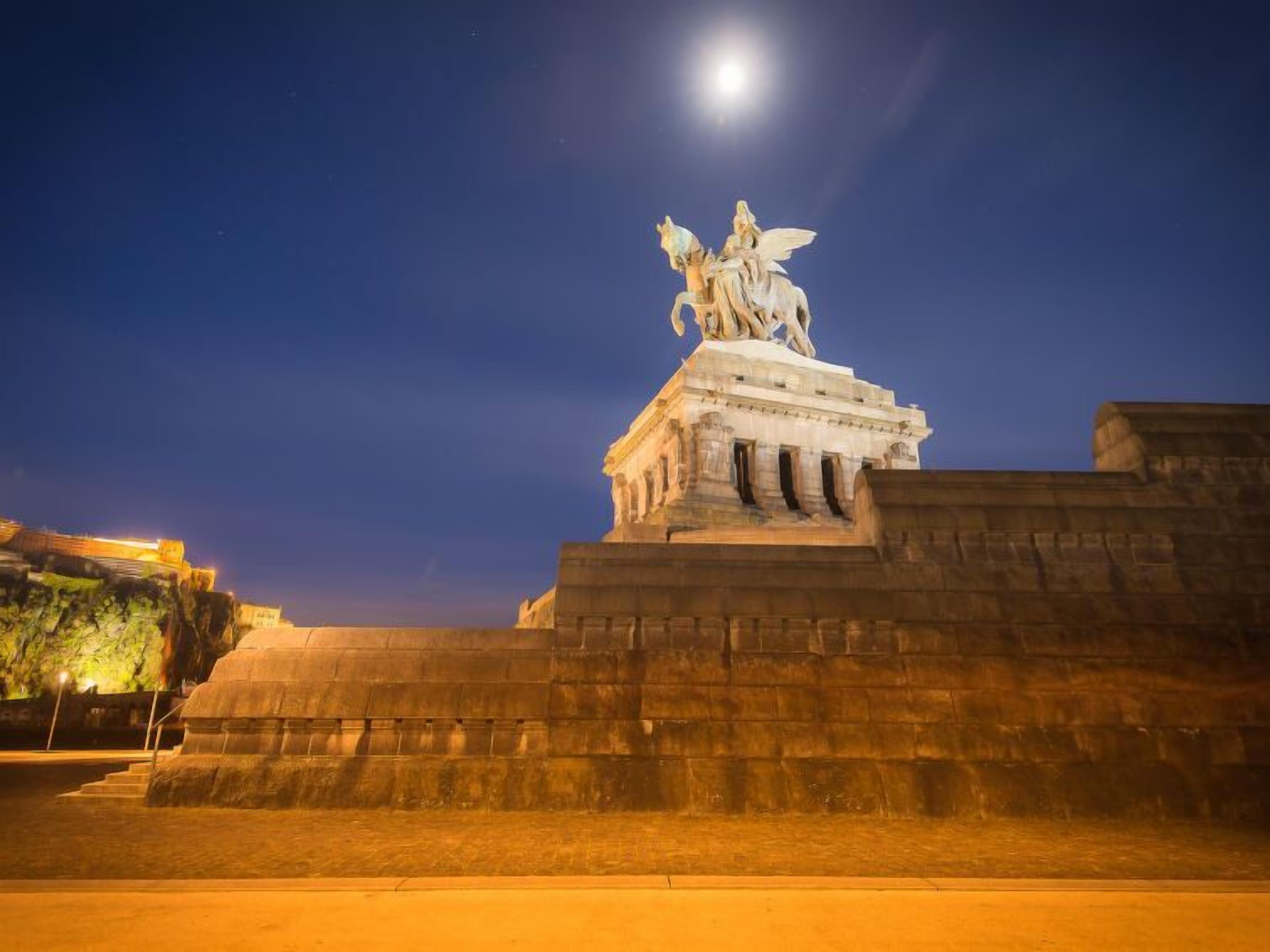 A nighttime view of a large, illuminated statue on a pedestal with a clear sky and moon in the background.