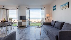 A modern living room with a large window overlooking a beach, a sofa, a coffee table, and a TV on a stand. - Les Caravelles by Interhome (Cabourg)