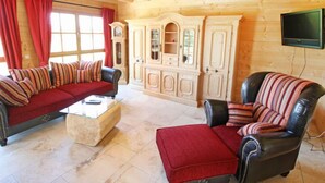 A cozy log cabin interior with a red sofa, a leather armchair, and a glass coffee table. A wooden cabinet with glass doors is against the wall, and a flat-screen TV is mounted above the armchair. - Apartment Lärche by Interhome (Grosserlach)