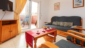 Een gezellige woonkamer met een bank, een houten tafel met kopjes, een televisie en een schuifpui naar een balkon.
