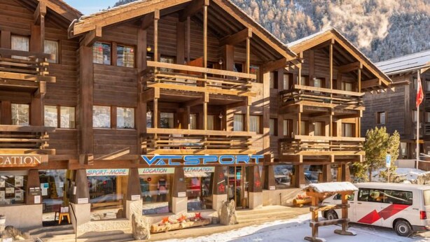 Un bâtiment en bois doté de balcons, une enseigne indiquant « VAL SPORT », et un paysage montagneux en arrière-plan.