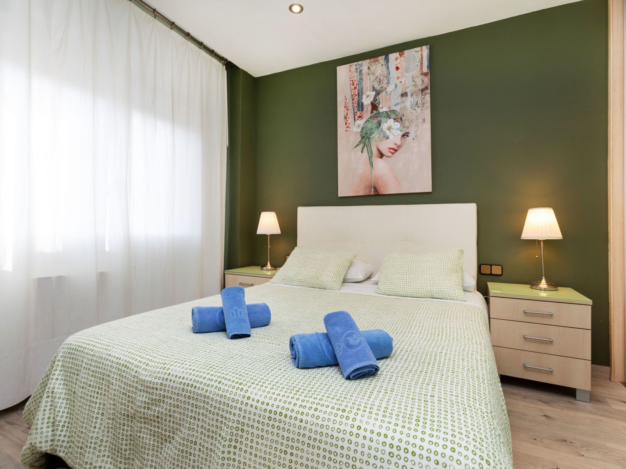 Un lit bien fait, recouvert d’une couvre-lit à motifs verts, avec deux serviettes bleues et un tableau représentant une femme entourée d’oiseaux, accroché au mur.