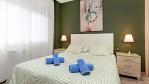 A neatly made bed with a green patterned bedspread, two blue towels, and a painting of a woman with birds on the wall.