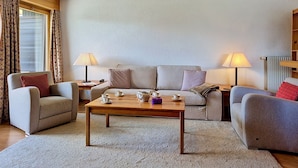 Een woonkamer met een bank, twee fauteuils, een salontafel, lampen en een raam met gordijnen.