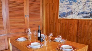 A wooden dining table set with plates, glasses, and cutlery, with a bottle of wine and a painting of a snowy mountain on the wall behind. - Les Moutières B1 et B2 (Val Claret) by Interhome (Tignes)