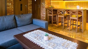 Een moderne woonkamer met een grijze bank, een houten salontafel met een kanttafelkleed, en een keukenhoek met houten kastjes en een barkruk.