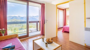 Ett hotellrum med en stor skjutdörr i glas som leder ut till en balkong med utsikt över bergen. Det finns en säng med ett rosa överkast, ett litet bord med en korg full av bröd och en vy över bergen genom fönstret.