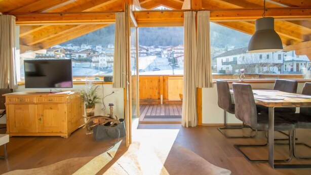 A spacious room with a wooden ceiling, a dining area with a table and chairs, a television on a wooden cabinet, and a view of a village through large windows. - Apartment Lanner by Interhome (Axams)