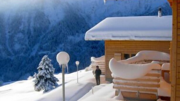 A wooden cabin with a snow-covered roof, surrounded by snow-covered mountains and trees.