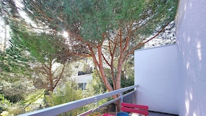 Un balcon avec une table rouge dressée pour deux personnes, entouré de verdure et d’arbres.