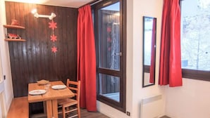 A room with a wooden table and chairs, a window with red curtains, and a wall-mounted shelf with decorative items. - Armoise 51 by Interhome (Les Menuires)