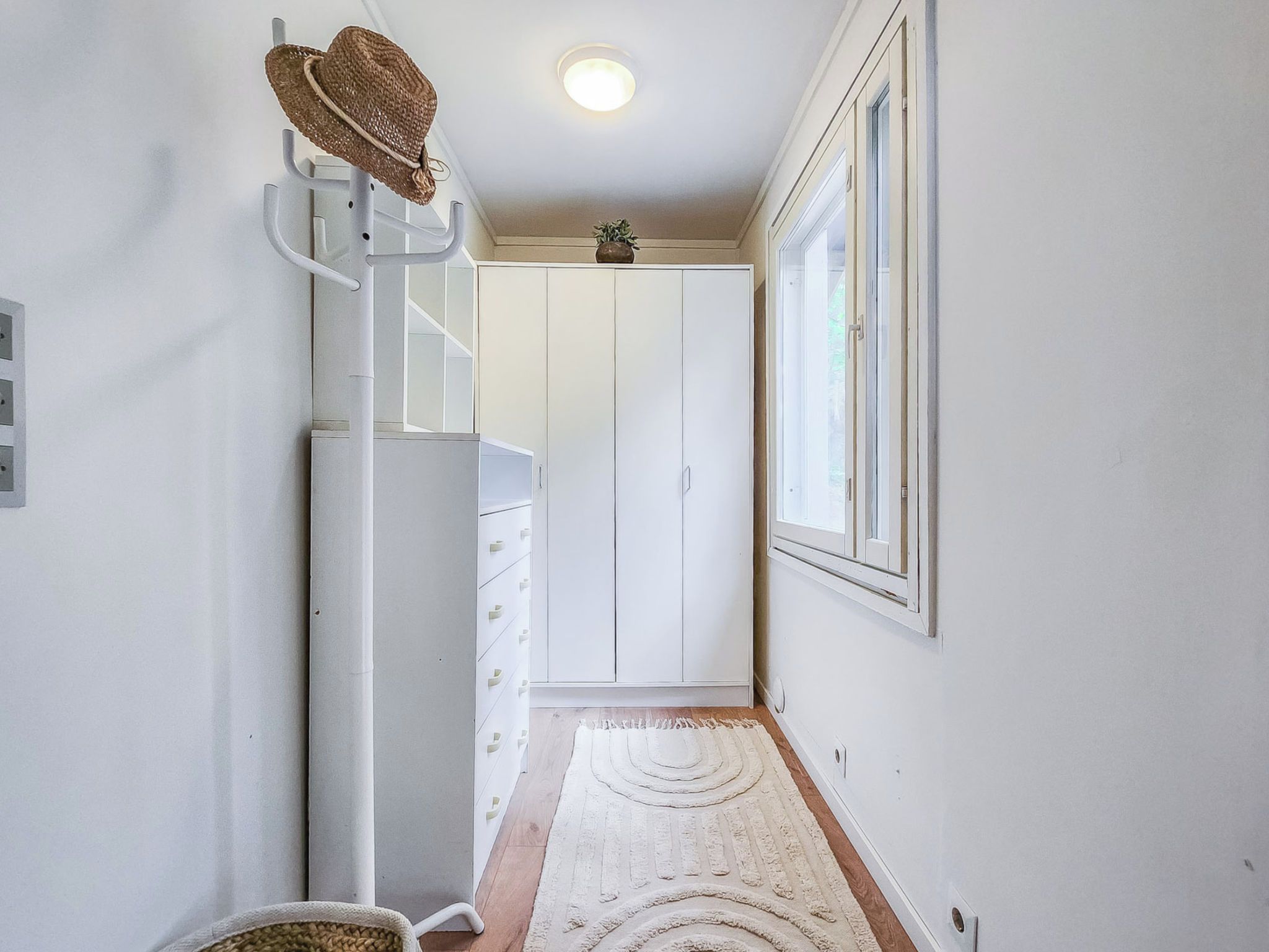 Ein Flur mit einem weißen Kleiderschrank, einem Haken für Hüte, einer kleinen Pflanze und einem Fenster mit Jalousien.
