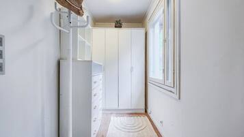 Ein Flur mit einem weißen Kleiderschrank, einem Haken für Hüte, einer kleinen Pflanze und einem Fenster mit Jalousien.
