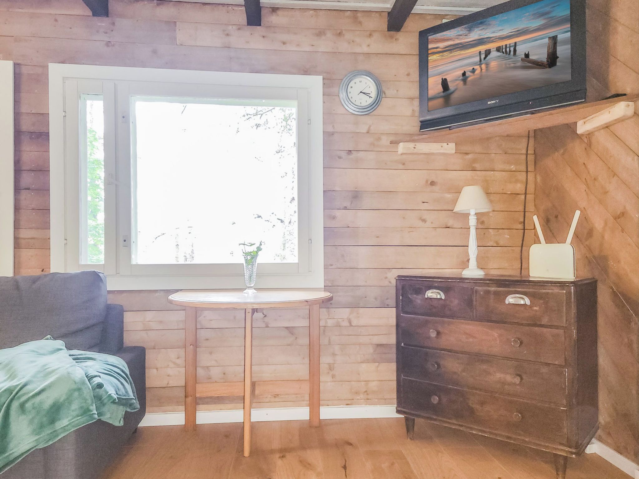 Een kamer met houten wanden, een raam, een bank, een klein tafeltje met een vaas, een dressoir met lades, een klok en een aan de muur bevestigde televisie.