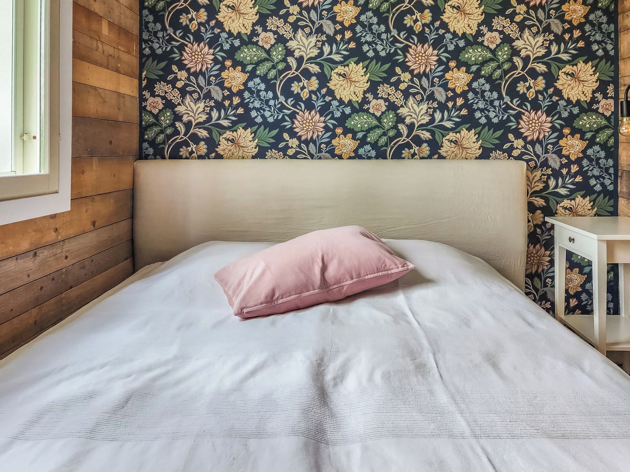 Un dormitorio con cabecera de madera, una cama individual con sábana blanca y almohada rosa, y un papel pintado floral.