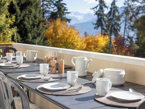 A table set for dining with white dishes, cups, and a teapot, overlooking a mountainous landscape with autumn foliage.