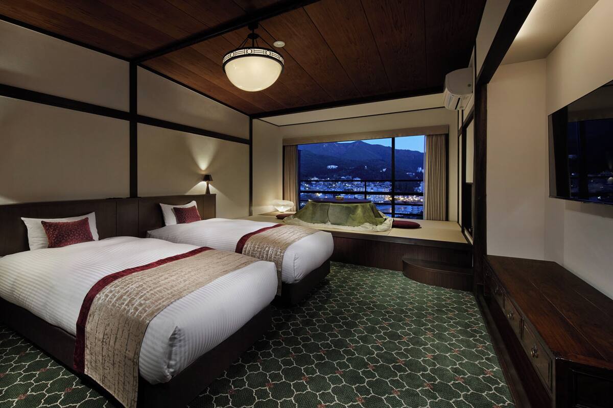 Ein Hotelzimmer mit zwei Betten, einem großen Fenster mit Blick auf die Stadt, einer Holzbalkendecke und einem grünen Teppich mit Muster.